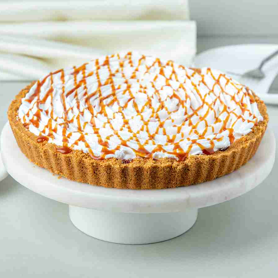 A baked banana cream pie topped with whipped cream, toasted coconut, and caramel sauce, served on a white plate with a drizzle of caramel sauce on the side.