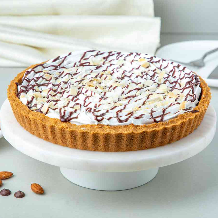 A Banoffee Pie on a white cake stand, topped with whipped cream, chocolate fudge, and roasted almonds, with a cookie crust and banana slices visible.