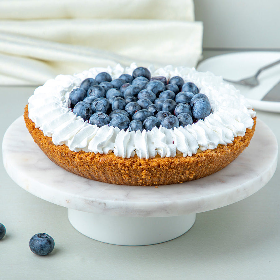 A blueberry cheesecake pie with fresh blueberries on top, presented on a white plate with a marble background.