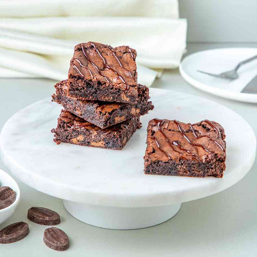 Stacked chocolate brownies on a white plate with chocolate chips scattered around, on a kitchen towel backdrop.