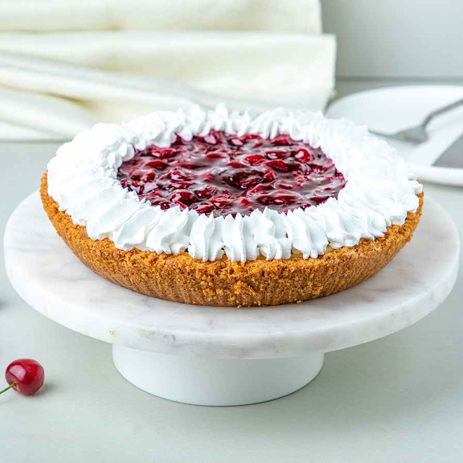 A freshly baked cherry cheesecake pie with a cookie crust, topped with sour cherry compote and whipped cream, on a white plate with cherries next to it.