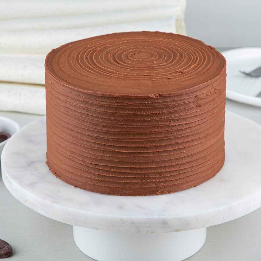 A chocolate ganache cake with chocolate buttercream frosting, placed on a marble surface, surrounded by chocolate pieces and a silver knife.