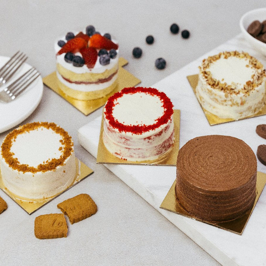 A variety of bento-style cakes with different toppings and fillings, including chocolate ganache, carrot cake, strawberry cream cake, honey lotus cake, and red velvet cake, presented on a table with a white background.