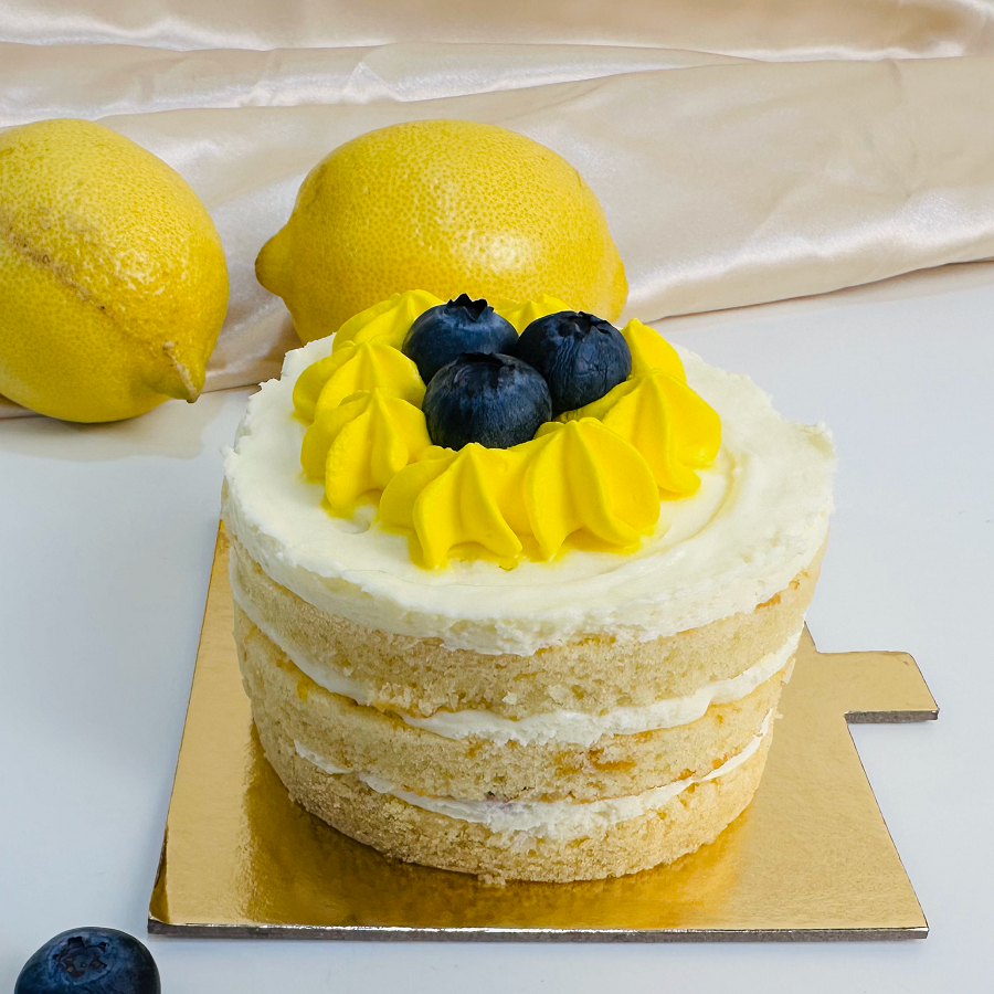 A small, round Lemon Blueberry Bento Cake with white frosting and blueberry decoration, served on a gold-colored plate with lemons and blueberries in the background.