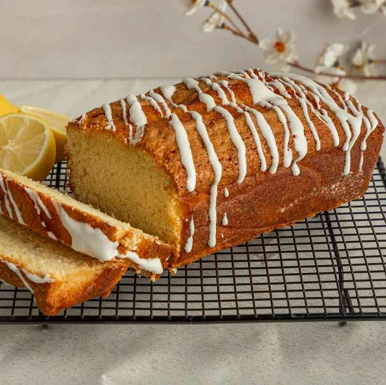 A loaf of lemon-glazed pound cake with a swirl of white frosting on top, placed on a cooling rack, with a slice cut out to show the layers, and a sprig of dill in the background.