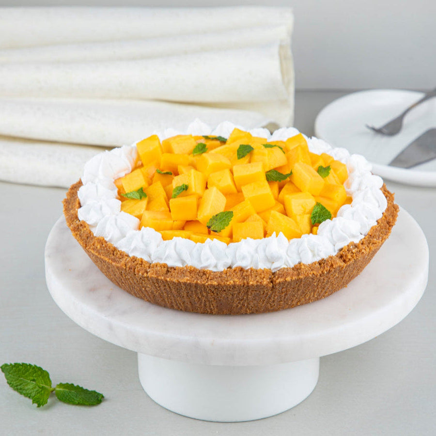 An 8-inch mango cheesecake pie with a cookie crust, topped with mango compote and fresh mango chunks, served on a white plate on a grey background.