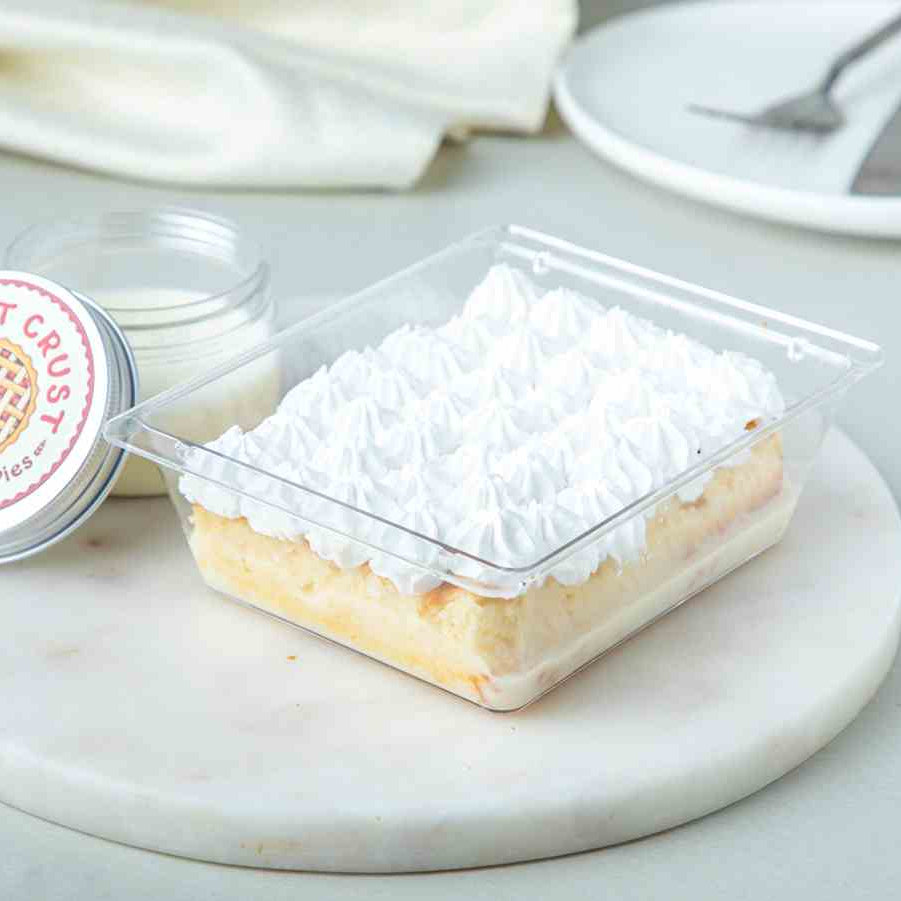 A Tres Leche cake with whipped cream on top, presented in a clear square container, with a label indicating it is the 'Original Milk Cake'.