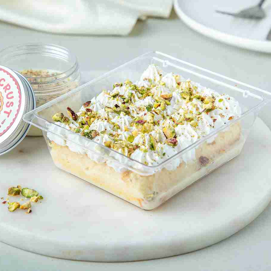 A packaged pistachio milk cake in a clear plastic container, topped with whipped cream and crushed pistachios, with a small jar of pistachio powder beside it.