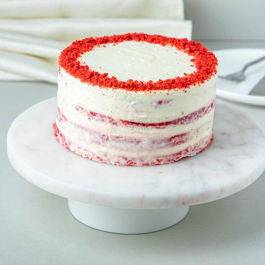 A red velvet cake with cream cheese frosting, garnished with red sprinkles around the base, on a white marble round cake stand.
