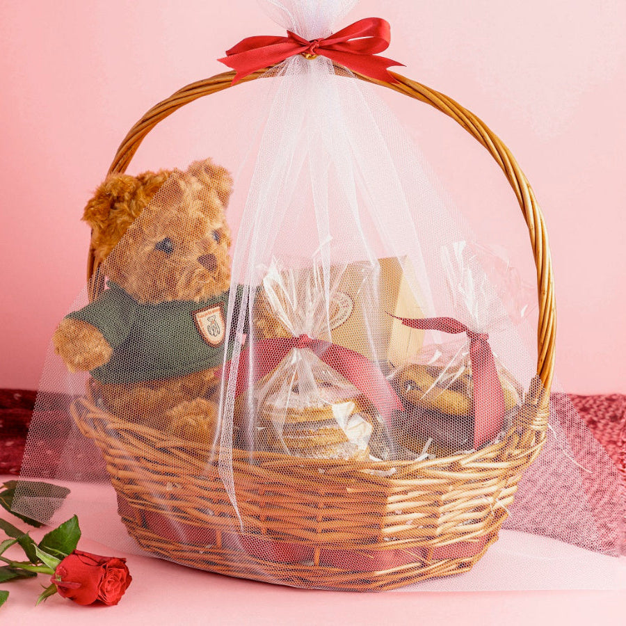 A Valentine's gift basket containing a soft toy, chocolate bento cake, cookies, and cherry cheesecake hand pies, all tied with white net and Ribbon.