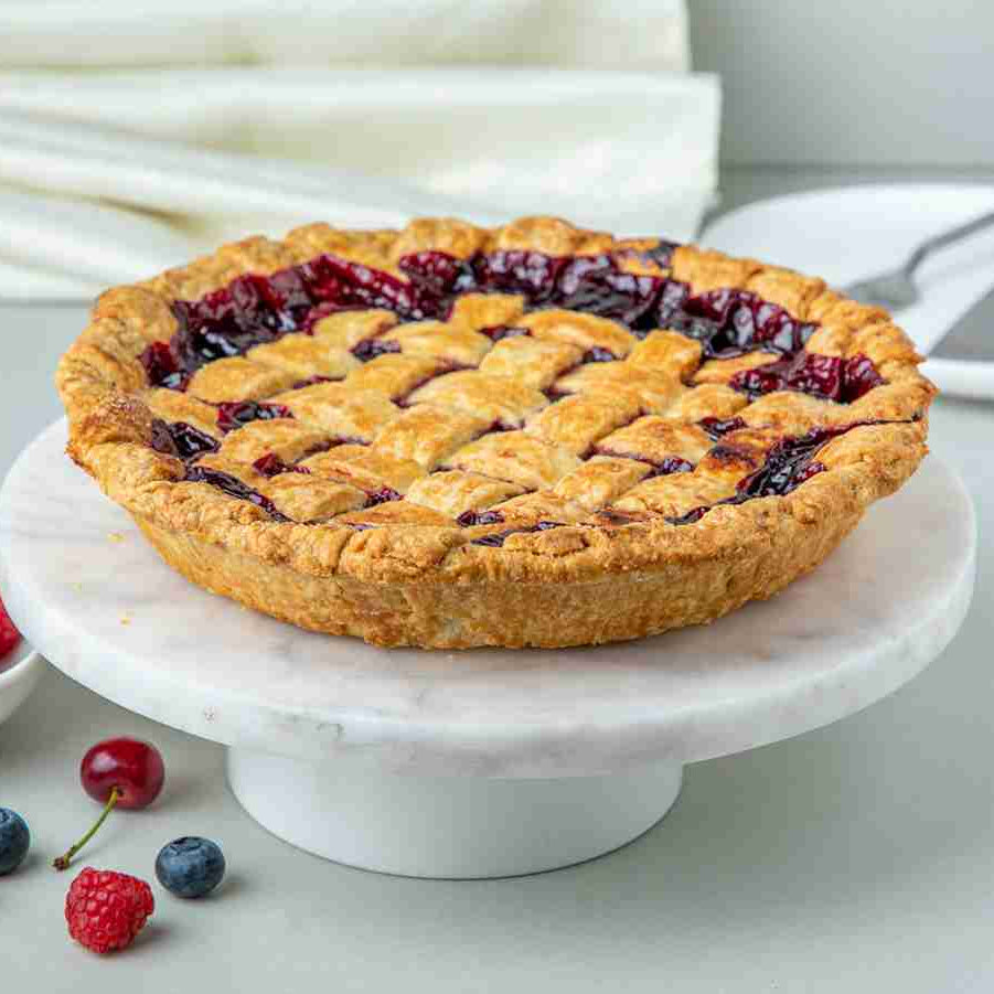 A baked pie with a lattice crust topped with a mix of blueberries, raspberries, strawberries, and cherries, on a white cake stand, with a bowl of berries beside it.
